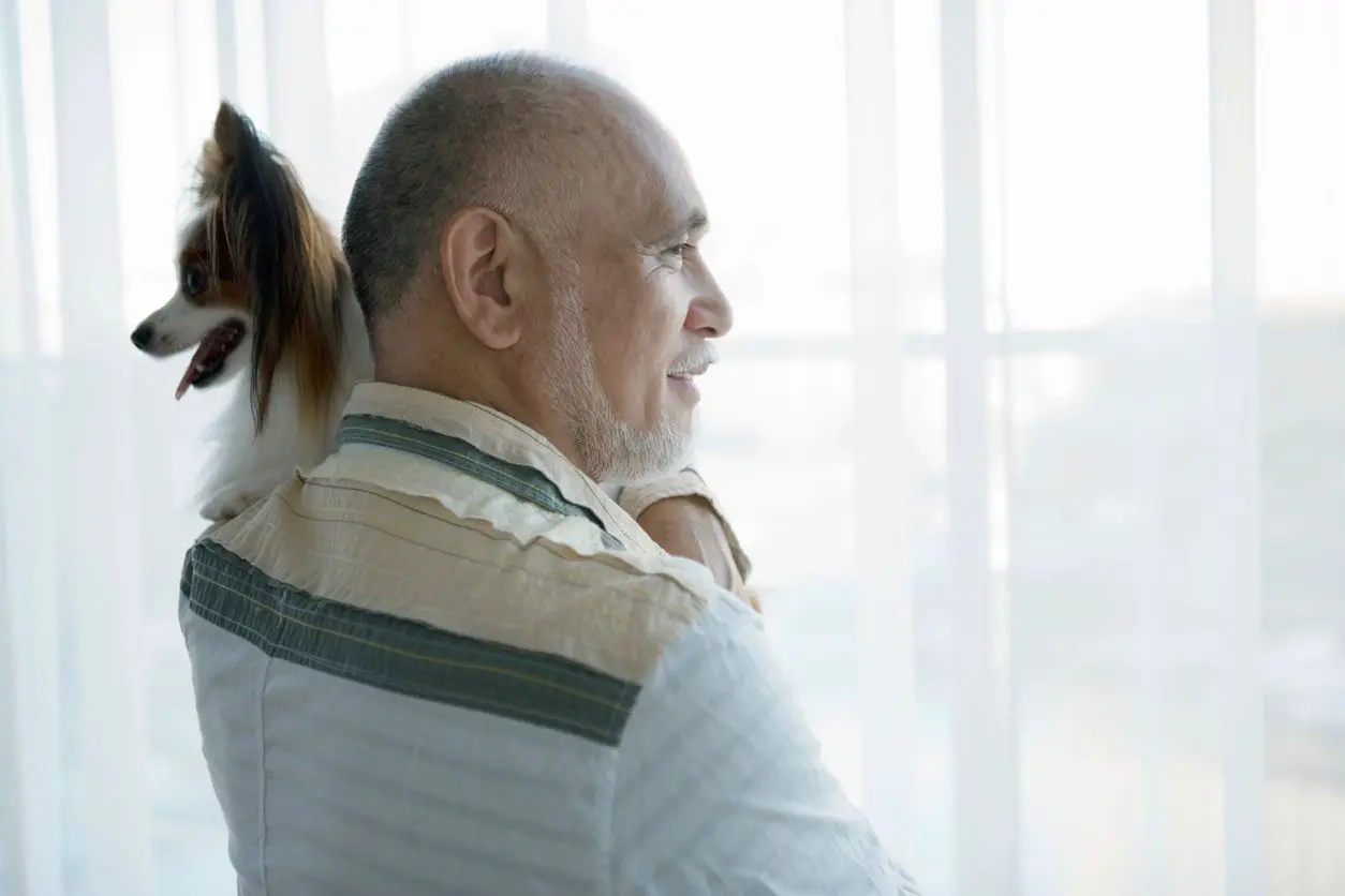 Senior man standing beside window, carrying dog,